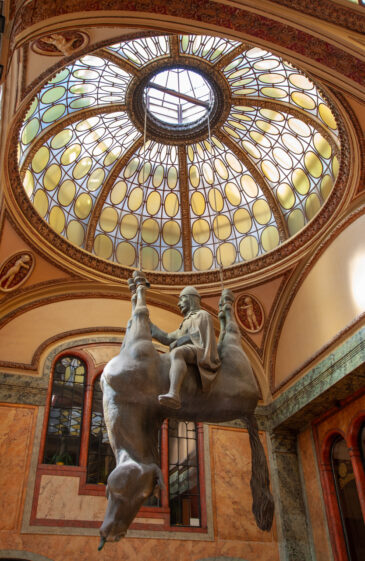 The upside-down horse statue in Prague is a satirical sculpture officially titled “Kůň” (Horse) by Czech artist David Černý. It hangs in the Lucerna Palace near Wenceslas Square, just a short walk from the statue of St. Wenceslas on horseback that it mocks. What it is: A parody of the famous St. Wenceslas statue nearby. Description: St. Wenceslas sits confidently atop a dead horse, which is hanging upside down by its legs, its tongue lolling out. Location: Lucerna Passage, a historic shopping arcade.