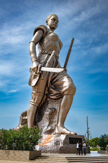 Standing at 30 metres, the Amazon Monument in Cotonou is an incredible tribute to the female warriors of Dahomey. With a rifle in hand and a defiant gaze, the Amazon embodies the strength, courage, and resilience of these legendary women.
Unveiled in 2022, the statue was commissioned by the Beninese government as part of an effort to reclaim and honor its pre-colonial history. It stands near the Place de l’Amazone, a key site in Cotonou’s ongoing cultural renaissance. It is meant as a statement: Benin’s past is not forgotten, and its female warriors remain a source of national pride.