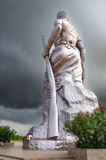 Standing at 30 metres, the Amazon Monument in Cotonou is an incredible tribute to the female warriors of Dahomey. With a rifle in hand and a defiant gaze, the Amazon embodies the strength, courage, and resilience of these legendary women.
Unveiled in 2022, the statue was commissioned by the Beninese government as part of an effort to reclaim and honor its pre-colonial history. It stands near the Place de l’Amazone, a key site in Cotonou’s ongoing cultural renaissance. It is meant as a statement: Benin’s past is not forgotten, and its female warriors remain a source of national pride.