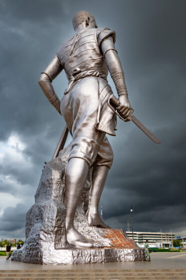 Standing at 30 metres, the Amazon Monument in Cotonou is an incredible tribute to the female warriors of Dahomey. With a rifle in hand and a defiant gaze, the Amazon embodies the strength, courage, and resilience of these legendary women.
Unveiled in 2022, the statue was commissioned by the Beninese government as part of an effort to reclaim and honor its pre-colonial history. It stands near the Place de l’Amazone, a key site in Cotonou’s ongoing cultural renaissance. It is meant as a statement: Benin’s past is not forgotten, and its female warriors remain a source of national pride.