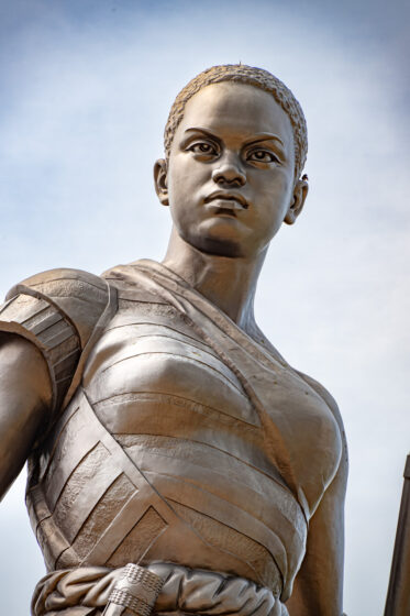 Standing at 30 metres, the Amazon Monument in Cotonou is an incredible tribute to the female warriors of Dahomey. With a rifle in hand and a defiant gaze, the Amazon embodies the strength, courage, and resilience of these legendary women.
Unveiled in 2022, the statue was commissioned by the Beninese government as part of an effort to reclaim and honor its pre-colonial history. It stands near the Place de l’Amazone, a key site in Cotonou’s ongoing cultural renaissance. It is meant as a statement: Benin’s past is not forgotten, and its female warriors remain a source of national pride.