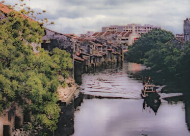 The Malacca River winds past rows of old, weathered shophouses built right to the water's edge, a small fishing boat making its way downstream toward the town centre.