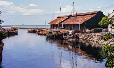 The Malacca River winds past rows of old, weathered shophouses built right to the water's edge, a small fishing boat making its way downstream toward the town centre.