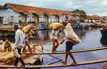 Dockworkers carry heavy sacks across planked walkways between boats at Malacca's harbour, 1978. Traditional wooden cargo vessels line the quay, with warehouses and a busy waterfront behind them.